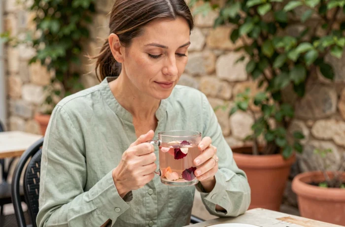 Femme tenant une tasse de thé floral à l'hibiscus et à la rose sur une terrasse de café ensoleillée, ambiance méditerranéenne apaisante